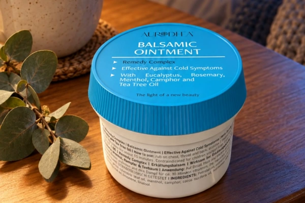 A jar of AURODHEA Balsamic Ointment with a turquoise lid on a wooden bedside table next to a cup of tea and a sprig of eucalyptus